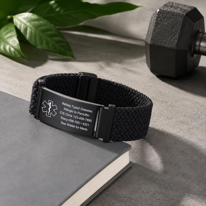 May include: A black medical alert bracelet featuring a woven nylon band and a rectangular metal plate. The plate displays the Star of Life symbol and engraved text: Kelsey, Type2 Diabetes, Allergic to Penicillin, ICE: Chris 123-456-7890, Stacy: 098-765-4321, See Wallet for Meds.