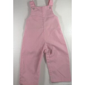 May include: A solid light pink overall featuring a high-waisted bib front with two shoulder straps. The garment includes two small white buttons at the top of the bib and a series of white snap closures along the inner leg seam for easy access.