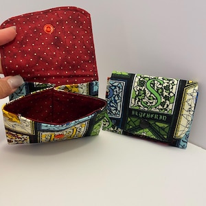 May include: Three fabric wallets. One is open, revealing a red interior with white polka dots. The other two wallets are closed, featuring a green and black pattern with the word "Slytherin". The wallets have a button closure.