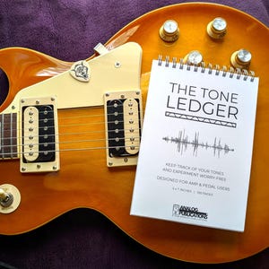 May include: A sunburst-finish electric guitar with a white notebook titled "THE TONE LEDGER" resting on its body. The notebook includes text about tracking tones and is designed for amp and pedal users. The guitar has gold knobs and black pickups.