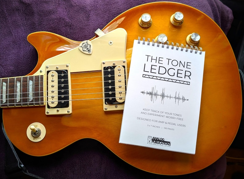 May include: A sunburst-finish electric guitar with a white notebook titled "THE TONE LEDGER" resting on its body. The notebook includes text about tracking tones and is designed for amp and pedal users. The guitar has gold knobs and black pickups.