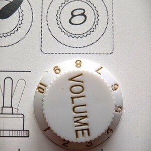May include: A white guitar volume knob with gold lettering and numbers. The word "VOLUME" is vertically centered. The numbers 6, 7, 8, 9, and 10 are visible. The image also shows a diagram with the number 8.
