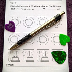 May include: A spiral-bound notebook with a silver pen and three guitar picks. The notebook has diagrams and text for guitar effects. The picks are purple, green, and dark green. The text includes "AMP PEDAL", "TONE", and "The Tone Ledger &copy; 2026".
