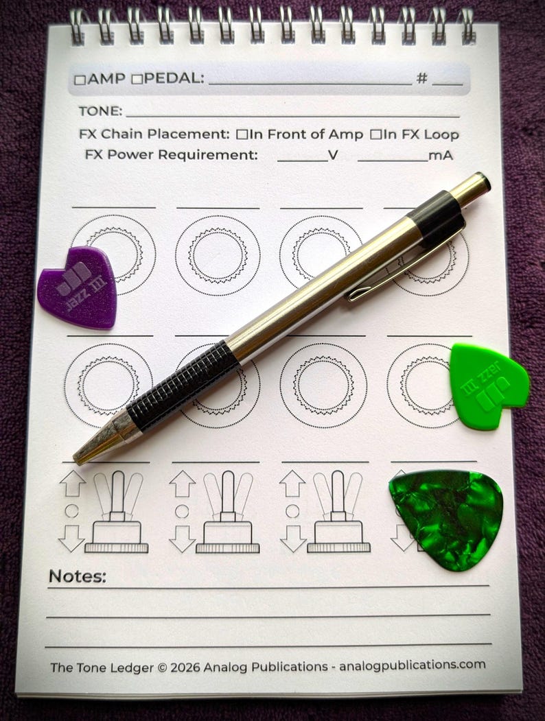 May include: A spiral-bound notebook with a silver pen and three guitar picks. The notebook has diagrams and text for guitar effects. The picks are purple, green, and dark green. The text includes "AMP PEDAL", "TONE", and "The Tone Ledger &copy; 2026".