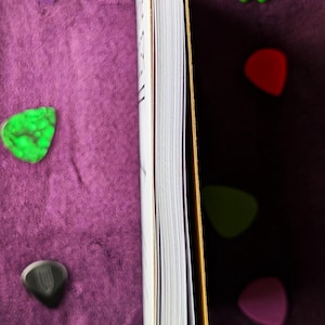 May include: A partially visible book with white pages and a gold spine, resting on a purple surface. Several colorful guitar picks in green, red, purple, and black are scattered around the book.