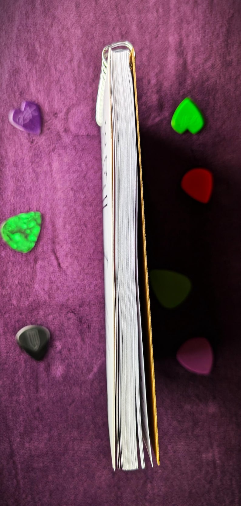May include: A partially visible book with white pages and a gold spine, resting on a purple surface. Several colorful guitar picks in green, red, purple, and black are scattered around the book.