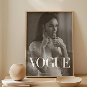 May include: A monochrome print of a woman holding a phone and lipstick, with the word "VOGUE" in large letters. The print is in a wooden frame, displayed on a light wood table with a beige vase and bowl.