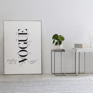 Puede incluir: Una impresi&oacute;n de Vogue en blanco y negro enmarcada se apoya contra una pared blanca. La impresi&oacute;n presenta el logotipo de Vogue en una disposici&oacute;n vertical con texto manuscrito. Dos mesas auxiliares con marcos de metal y una peque&ntilde;a planta est&aacute;n al lado de la impresi&oacute;n.