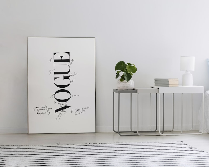 Puede incluir: Una impresi&oacute;n de Vogue en blanco y negro enmarcada se apoya contra una pared blanca. La impresi&oacute;n presenta el logotipo de Vogue en una disposici&oacute;n vertical con texto manuscrito. Dos mesas auxiliares con marcos de metal y una peque&ntilde;a planta est&aacute;n al lado de la impresi&oacute;n.