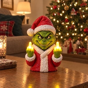 Op de afbeelding: Een groen, getextureerd karakterfiguur met een rode kerstmuts en jas met witte imitatiebontrand. Het figuur heeft een ondeugende gezichtsuitdrukking en steekt beide middelvingers op, die verlicht zijn met heldergele LED-lampjes.