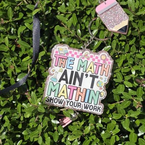 Peut inclure: Un porte-badge carré avec une finition pailletée et le texte "THE MATH AIN'T MATHIN' SHOW YOUR WORK" accompagné d'icônes scolaires colorées. Il comprend une breloque en forme de crayon, un cordon noir et un pompon rose clair.