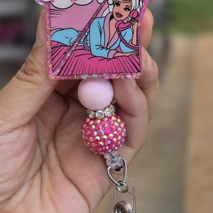 May include: A badge reel featuring a rectangular resin topper with a comic-style illustration of a person in a robe and towel talking on a pink phone, with the text "Me trying to figure out who you're talking to...". It has pink and glittery beads.