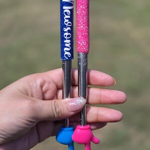 May include: Two extendable pointers with glittery handles, one blue and one pink. The blue handle features the white text "Mrs. Newsome". Both pointers have metallic shafts and end in small, solid-colored hand-shaped tips in blue and pink.