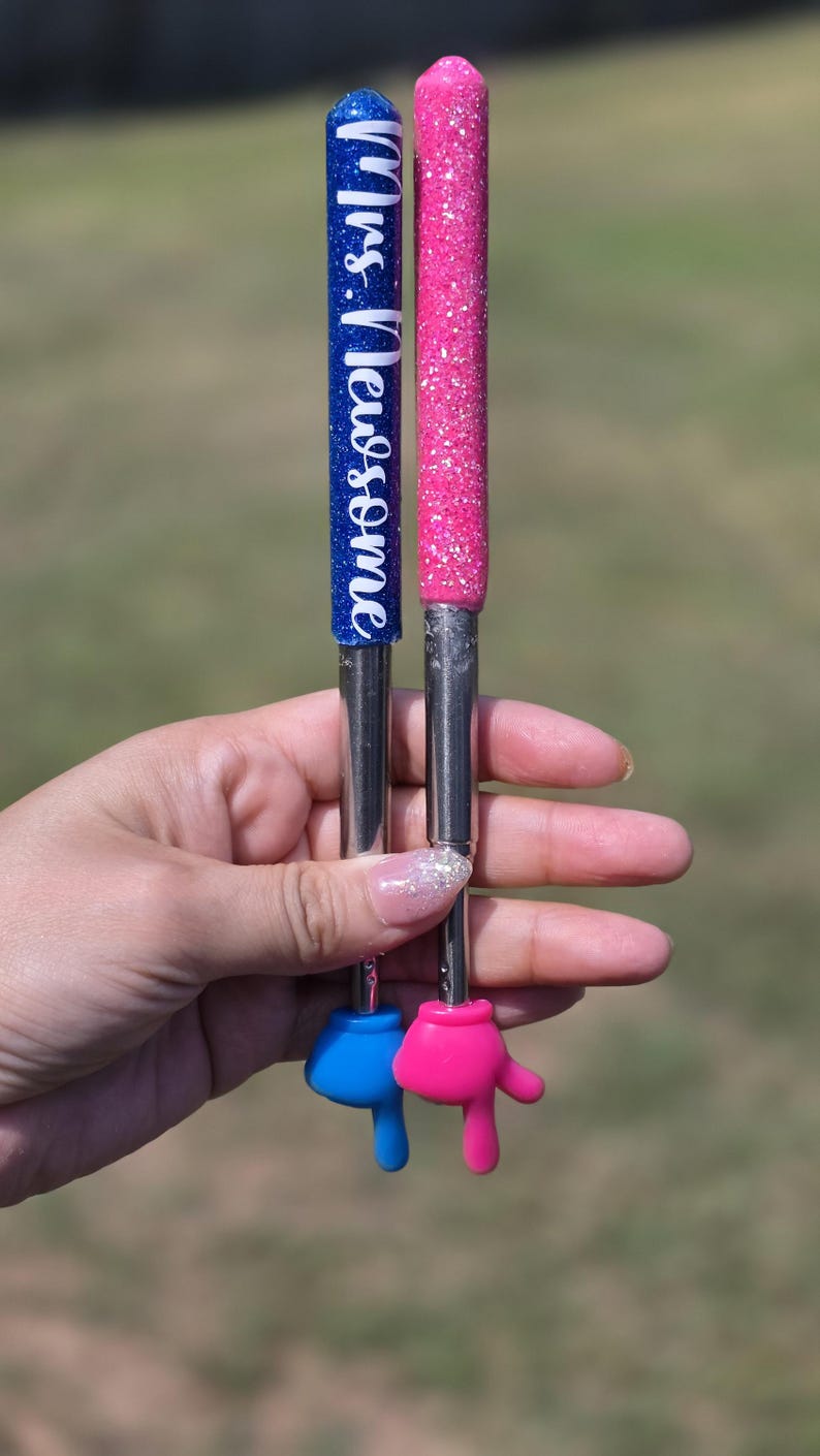 May include: Two extendable pointers with glittery handles, one blue and one pink. The blue handle features the white text "Mrs. Newsome". Both pointers have metallic shafts and end in small, solid-colored hand-shaped tips in blue and pink.