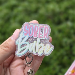 May include: A glittery badge reel featuring the text SOBER BABE in pink and light blue lettering. The badge reel has a clear, sparkly acrylic topper with a heart-shaped attachment connecting the reel to the decorative piece.