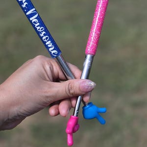 May include: Two pens with glittery barrels, one blue with the white text Mrs. Newsome and one pink. Each pen features a metallic silver shaft and a small plastic hand at the tip, with one hand colored pink pointing down and one blue hand making a peace sign.