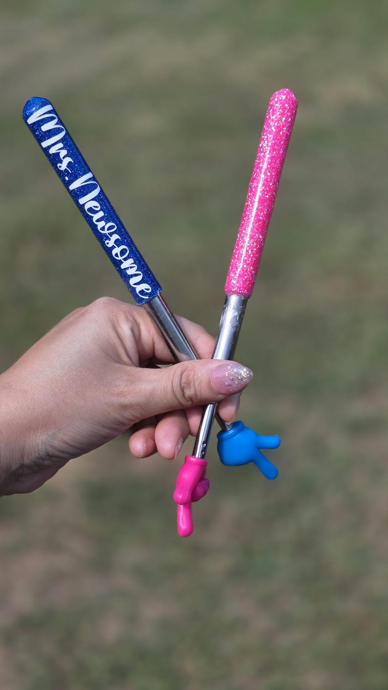 May include: Two pens with glittery barrels, one blue with the white text Mrs. Newsome and one pink. Each pen features a metallic silver shaft and a small plastic hand at the tip, with one hand colored pink pointing down and one blue hand making a peace sign.