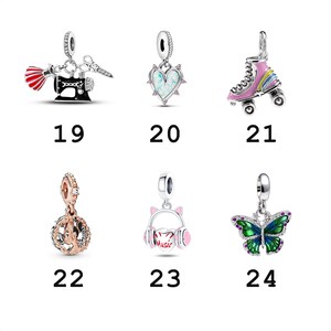 May include: A collection of six silver-toned charms labeled 19 through 24. The charms feature a sewing machine with scissors, a heart with iridescent inlay, a pink roller skate, a rose-gold filigree sphere, cat-ear headphones, and a butterfly.