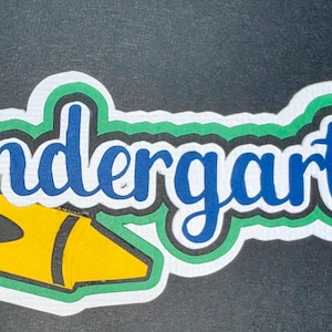 Könnte beinhalten: Ein farbenfroher Kindergarten-Aufkleber mit einem gelben Buntstift und einem roten Apfel. Das Wort "Kindergarten" ist in blauer Schrift mit weißer und grüner Umrandung geschrieben. Der Aufkleber ist für Bildungszwecke bestimmt.