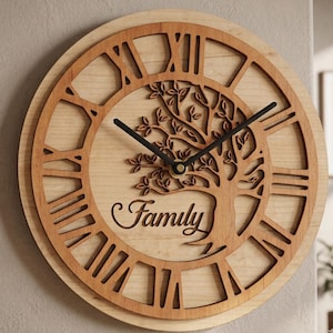 May include: Wooden wall clock with a light wood grain background and a darker wood frame. The clock features Roman numerals, black clock hands, and a tree design with the word "Family" in cursive.