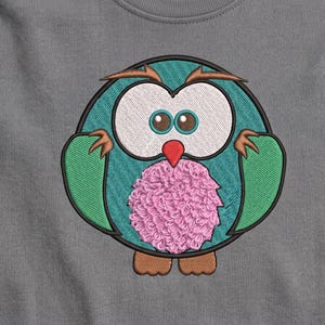 Puede incluir: Una camiseta gris con un diseño circular bordado de un búho. El búho tiene cuerpo azul cerceta, grandes ojos blancos con pupilas marrones, alas verdes, pico rojo, patas marrones y una barriga rosa texturizada. El bordado está delineado en negro.