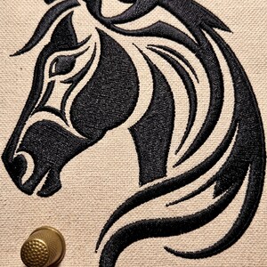 May include: A black embroidered horse head design on a light beige fabric background, featuring a stylized mane and facial contours. A small, metallic gold-colored thimble is positioned in the lower left corner of the textile surface.
