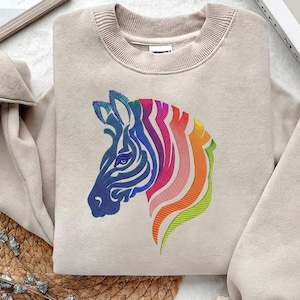 Może przedstawiać: Beżowa bluza z haftowaną głową zebry. Głowa zebry jest niebieska, a grzywa w kolorach tęczy. Bluza ma okrągły dekolt i długie rękawy. Kolorowy i przyciągający wzrok design.