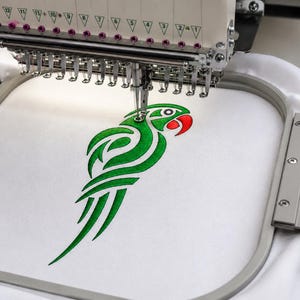 May include: An embroidery machine in operation, stitching a green parrot design with a red beak onto white fabric. The machine's needle is in action, creating the detailed artwork. The design is framed within an embroidery hoop.