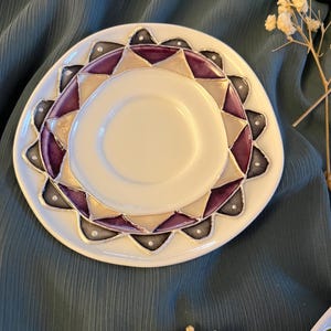 May include: A white ceramic saucer featuring a decorative border of alternating purple, beige, and dark gray triangular shapes. The design is outlined in silver metallic lines, with small white dots accenting the points of the outer pattern.