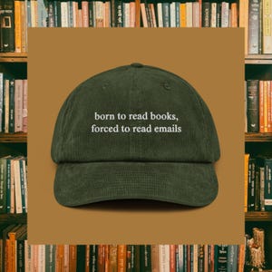 May include: Dark green corduroy baseball cap with the white text "born to read books, forced to read emails". The hat has a curved peak and a classic six-panel design. A bookshelf is in the background.