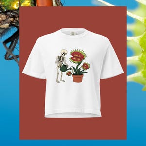 May include: A white short-sleeve t-shirt featuring a graphic print of a human skeleton holding a dark green watering can. The skeleton is watering a Venus flytrap plant with red and green leaves, which is potted in a brown terracotta pot.