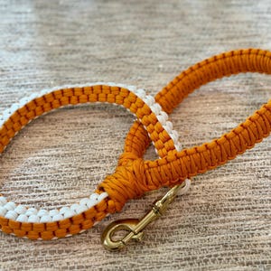 Op de afbeelding: Een gevlochten hondenriem in oranje en wit, met een messingkleurige sluiting. De riem is gemaakt van geweven materiaal, met een lus handvat en een contrasterende witte sectie.