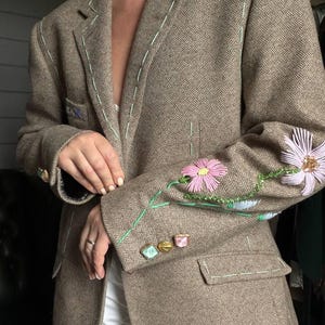 以下が含まれることがあります： 袖に花の刺繍が施された茶色のヘリンボーン柄ブレザー。ブレザーは、薄緑色の装飾的なステッチとカラフルなボタンが特徴です。花はピンクとラベンダーです。