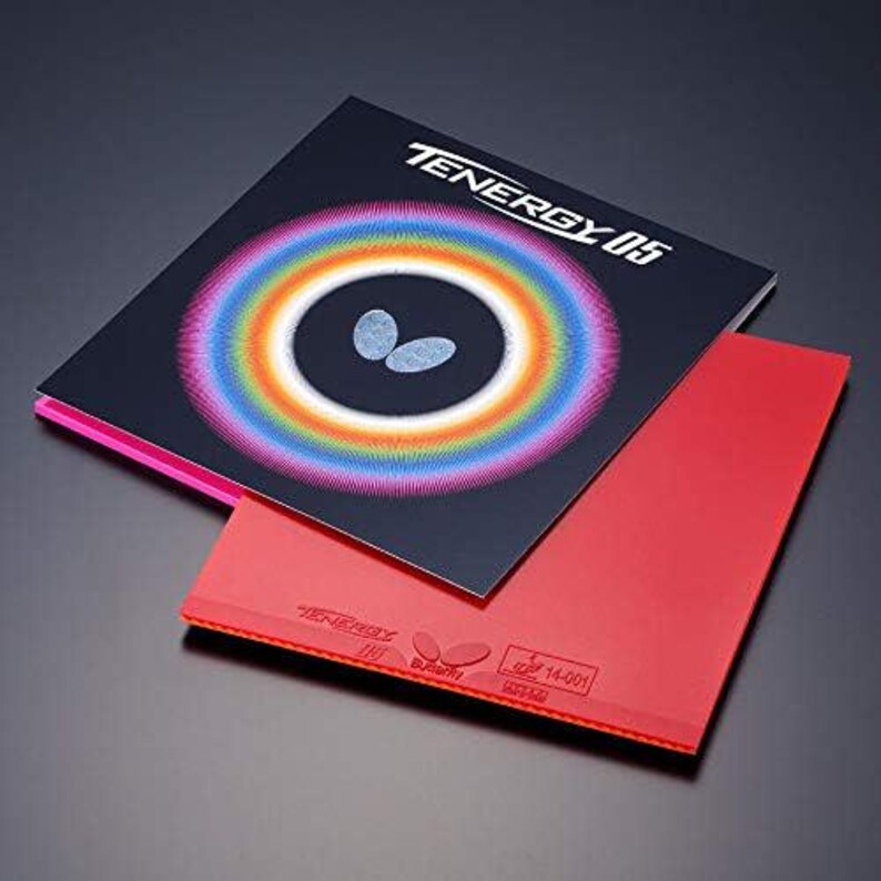May include: A red table tennis rubber sheet with a textured surface, featuring the embossed text TENERGY 05 and the Butterfly logo. It is partially covered by a black square packaging sleeve with a rainbow-colored circular graphic and text.