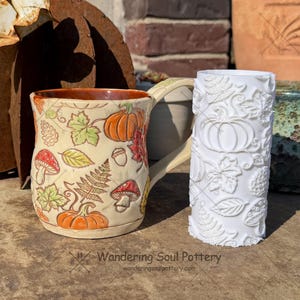 May include: A ceramic mug with a cream-colored glaze featuring hand-painted orange pumpkins, red mushrooms, and green leaves, paired with a white cylindrical texture roller embossed with matching pumpkin, leaf, and mushroom patterns.