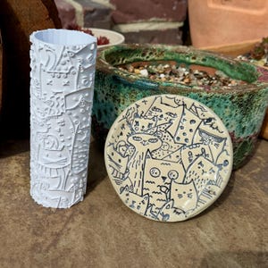 May include: A white cylindrical roller with a raised cat pattern and a circular beige ceramic dish featuring a black line art illustration of multiple cats. The roller and dish are positioned together, showcasing detailed feline motifs.