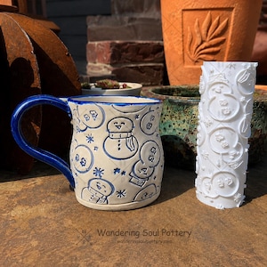May include: A cream-colored ceramic mug with a blue handle and a blue-outlined embossed pattern of snowmen and snowflakes, accompanied by a white cylindrical clay texture roller featuring a matching embossed snowman and snowflake design.