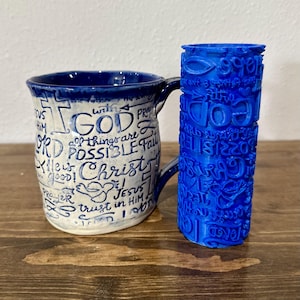 May include: A ceramic mug with a blue rim and handle, featuring handwritten script with the words "GOD", "Christ", and "Jesus". Beside it, a blue cylindrical object with similar text.