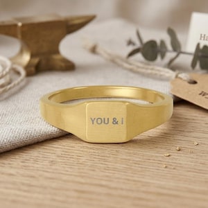 Könnte beinhalten: Ein goldfarbener Siegelring mit der Aufschrift "YOU & I" auf einer quadratischen Fläche. Der Ring liegt auf einer Holzoberfläche, zusammen mit einem Anhänger mit der Aufschrift "Handmade with Love".