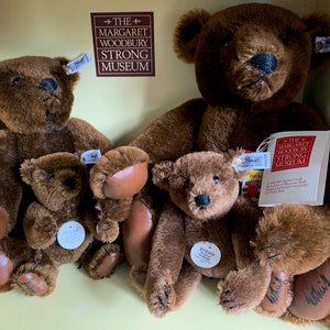 May include: Four brown teddy bears of different sizes, all with black eyes and noses. The largest bear is sitting with its paws outstretched. The other three bears are sitting in front of the largest bear. The bears are all wearing tags that say "Steiff" and "The Margaret Woodbury Strong Museum".