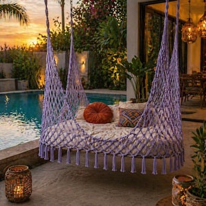May include: A lavender macrame swing bed with a net design, tassels, and a cream mattress. It has orange and patterned cushions. The swing is suspended by chains, set against a backdrop of a pool and sunset.