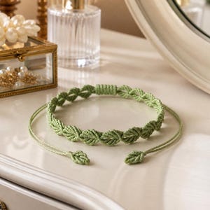 May include: A light green macrame bracelet with a leaf design. The bracelet is on a white surface, with a gold-trimmed glass jewelry box and a perfume bottle in the background. The bracelet has a leaf pattern and adjustable ties.