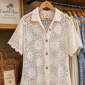 May include: A cream-coloured, short-sleeved crochet shirt with a collared neckline and wooden buttons. The shirt is displayed on a wooden hanger. The sign reads "Coastal Muse Resort Wear Made for Sunshine".