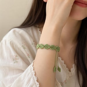 Puede incluir: Una pulsera de macramé verde con un diseño de hojas. La pulsera está hecha de cordón verde tejido y tiene una estética delicada y natural. La pulsera es ajustable y tiene dos pequeños dijes de hojas colgando.