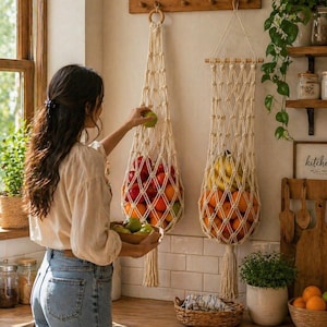 May include: Two macrame fruit holders, one filled with red apples and the other with oranges and bananas, hang on a wall. A person is holding a bowl of green apples. The kitchen has wooden shelves and a wooden countertop.