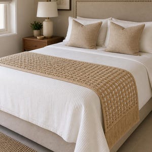 Può includere: Una scena di camera da letto con un letto con lenzuola bianche e due cuscini beige. Un copriletto beige intrecciato con un bordo a frange è posto sul letto. Un comodino in legno, una lampada e opere d'arte completano la stanza.
