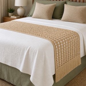 May include: A bed featuring a white duvet, olive green valance, and a tan crochet bed runner with fringe. Two tan and two olive green pillows are on the bed. A wooden headboard and framed picture are visible.