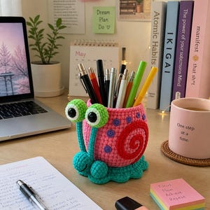 Könnte beinhalten: Ein handgefertigter, rosa gehäkelter Stifthalter in Schneckenform mit grünen Augen und einem roten Spiralmuster. Auf dem Schreibtisch befinden sich auch ein Laptop, Bücher, ein Notizblock, Stifte und Haftnotizen mit dem Text "Focus Plan Achieve Repeat".