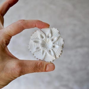 May include: A white, intricately carved, circular object with a floral and geometric design. The object is held between fingers, showcasing its detailed patterns and textures. The design features a central flower-like shape surrounded by radiating lines and dotted accents.