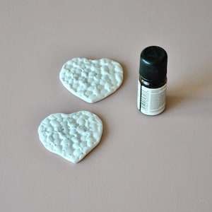 Puede incluir: Dos decoraciones blancas en forma de corazón texturizado y una pequeña botella de aceite esencial con tapa negra. La botella tiene una etiqueta blanca con texto. Dispuestas sobre una superficie rosa claro.